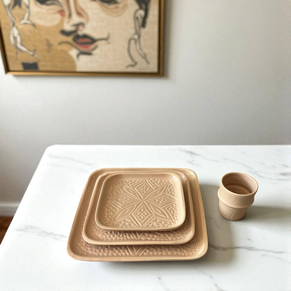 Set of ceramic trays and a cup on a marble surface with a wall art piece in the background.