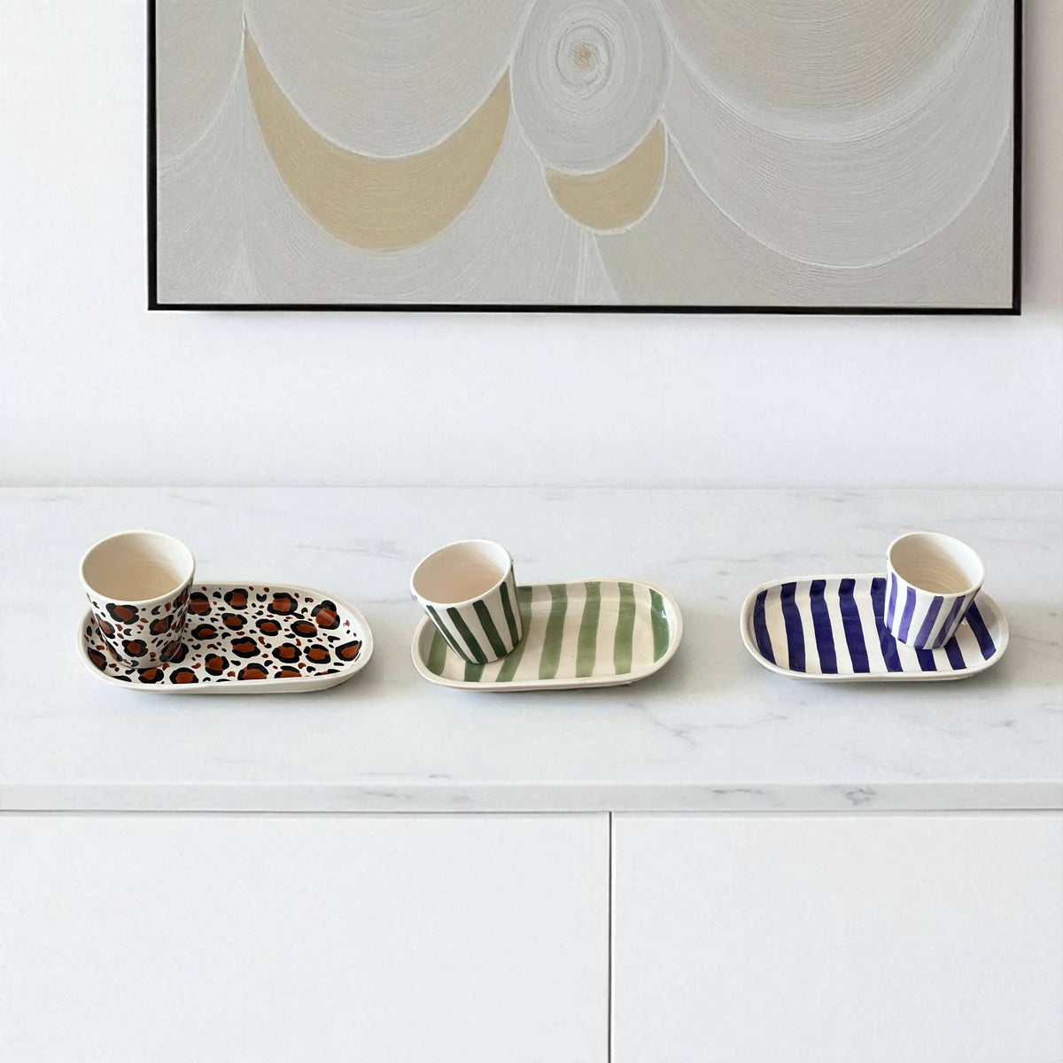 Set of ceramic cups and saucers with patterned designs on a white surface, framed abstract art in the background.