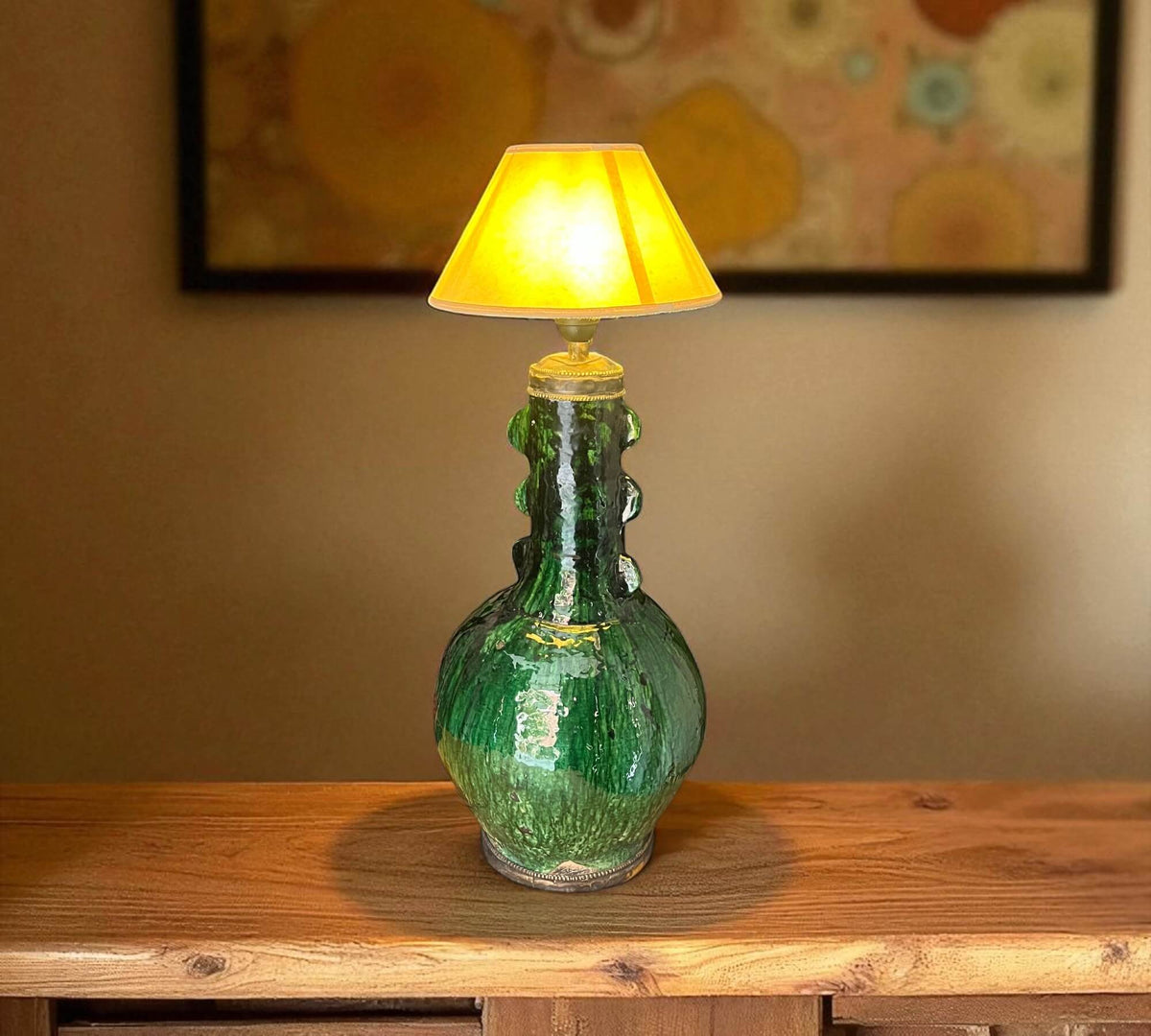 Green ceramic lamp with a yellow lampshade on a wooden surface, with a blurred abstract painting in the background.