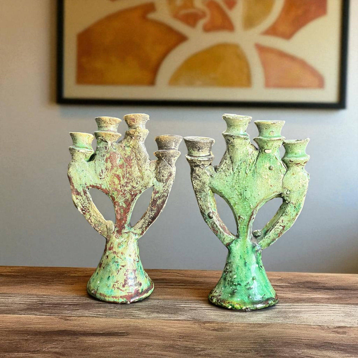 Two green ceramic candle holders on a wooden surface with a framed abstract painting in the background.