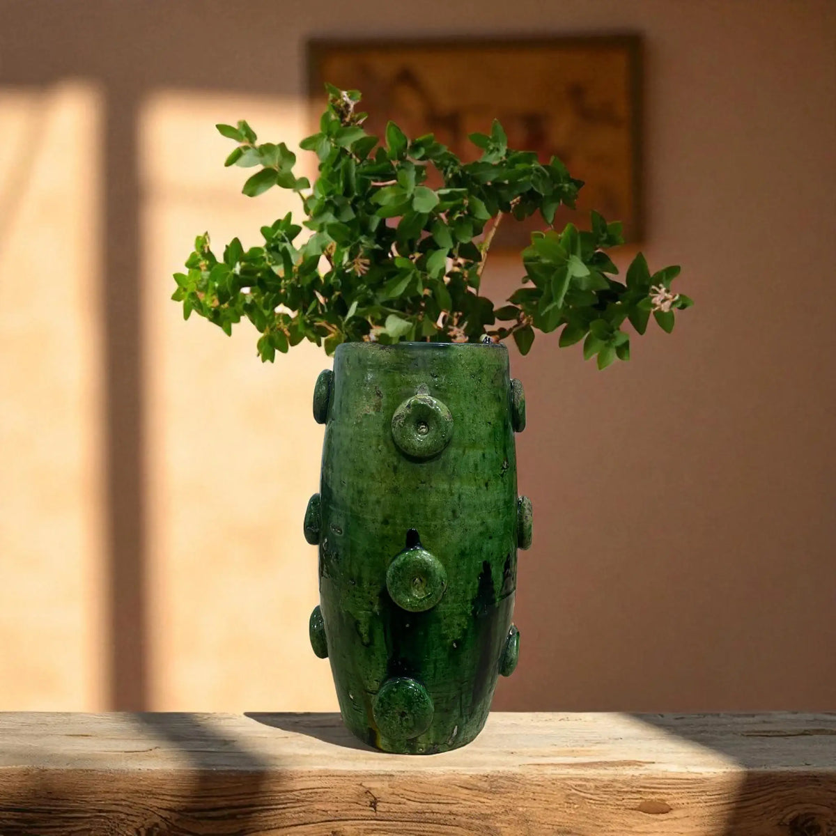 Green ceramic vase with plant on a wooden surface against a beige wall