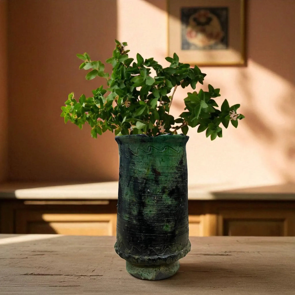 Green vase with a plant on a wooden table in a softly lit room.