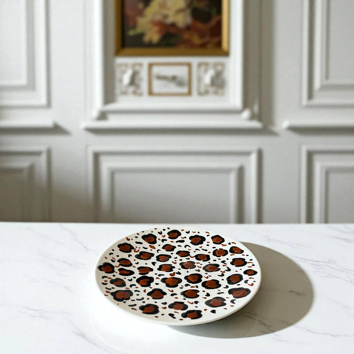 Round ceramic plate with leopard print design on a white surface, with a neutral wall and framed picture in the background.