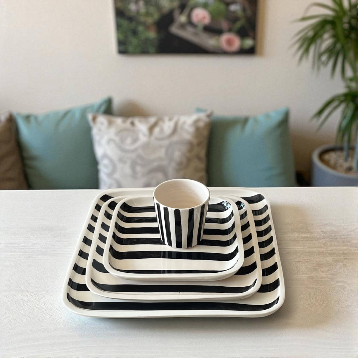 Set of black and white striped ceramic trays and cup on a table with a couch and plant in the background.