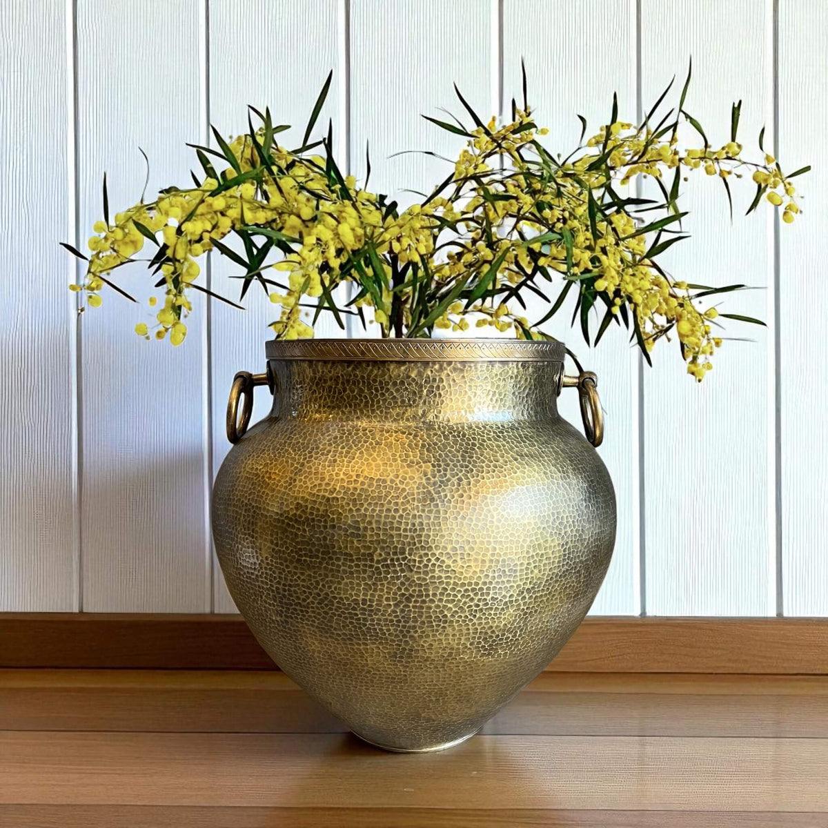 Decorative brass vase with yellow flowers on a wooden surface against a white paneled wall.