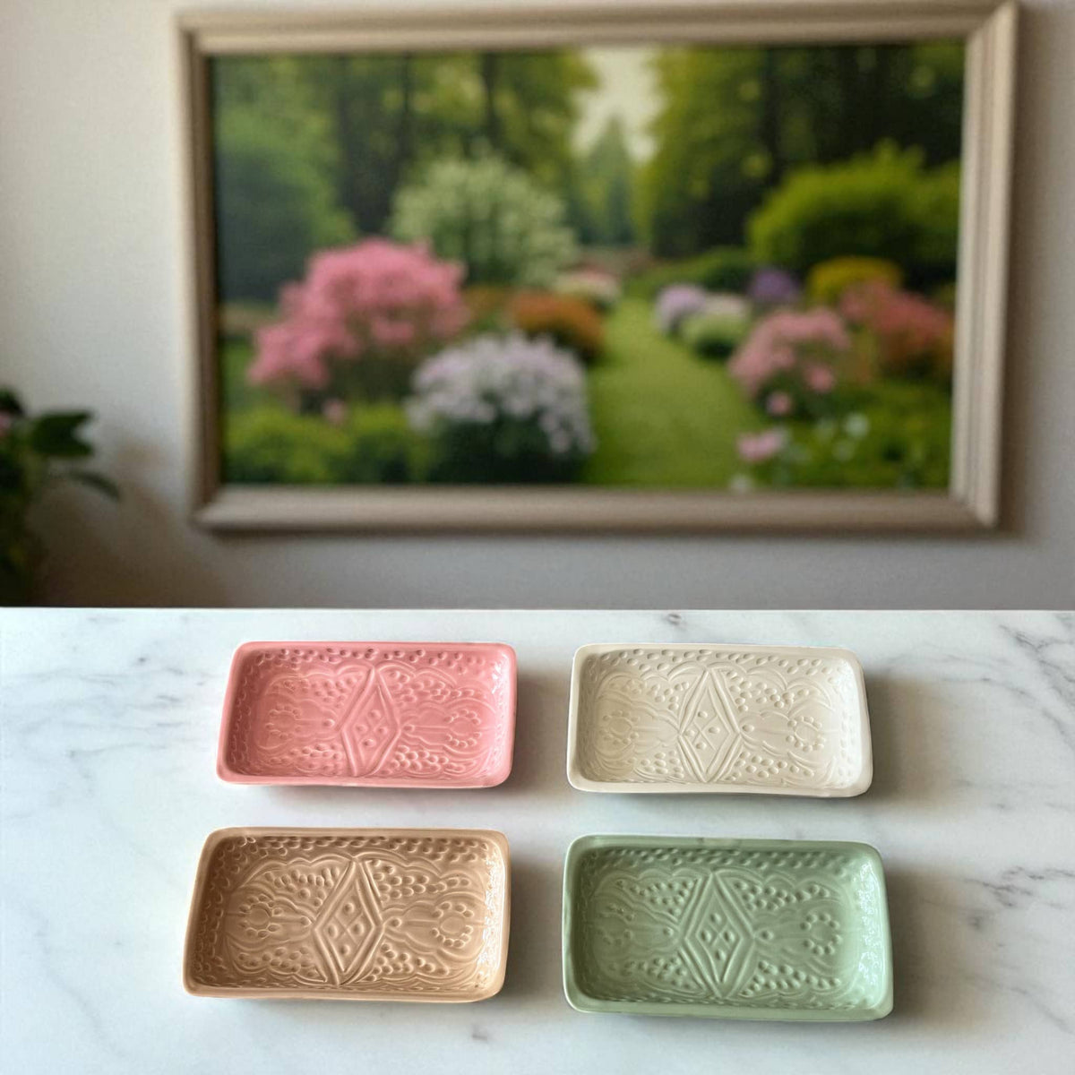 Four ceramic trays in pink, beige, gold, and green on a marble surface with a framed picture of a garden in the background.