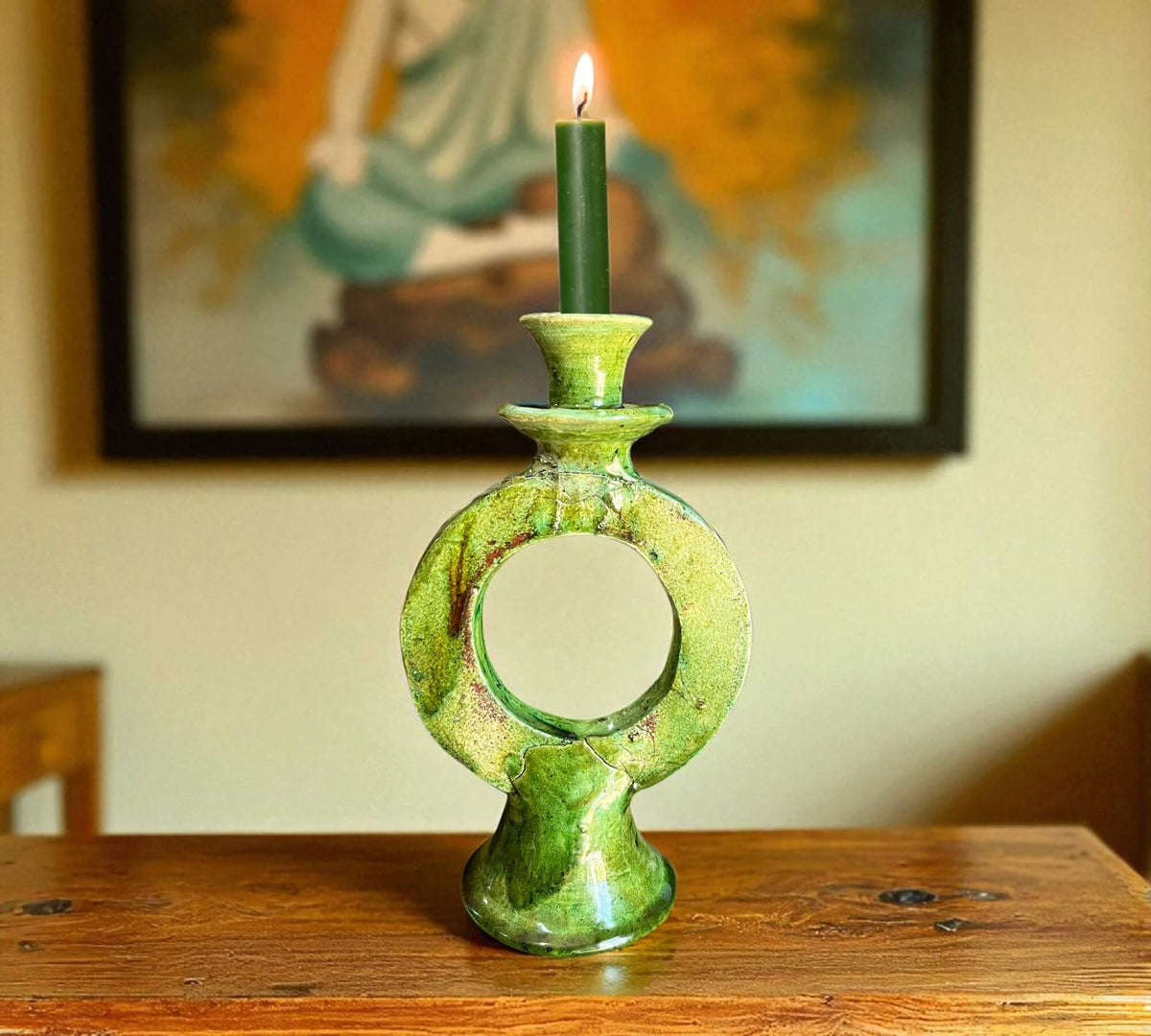 Green ceramic candle holder with a lit candle on a wooden surface, with a blurred painting in the background.