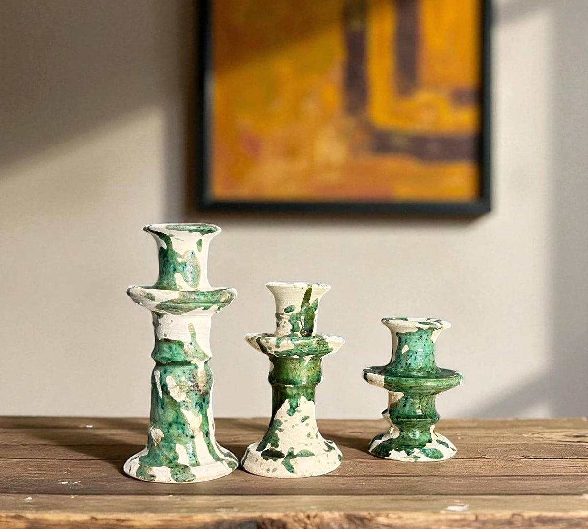 Three green and white ceramic candle holders on a wooden surface with a blurred abstract painting in the background.