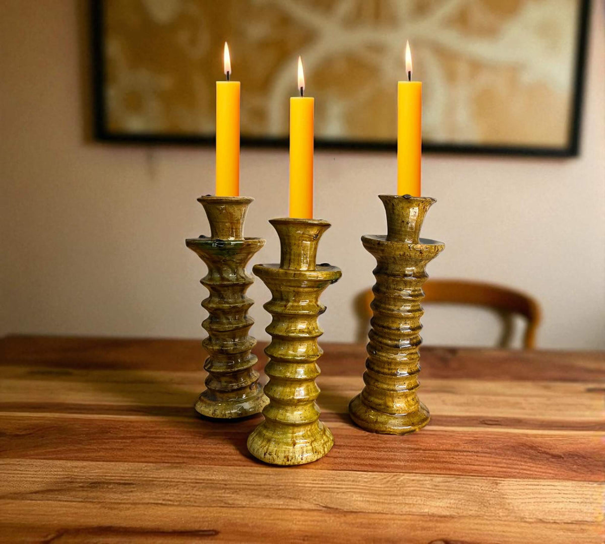 Three candle holders with lit candles on a wooden surface, framed artwork in the background.