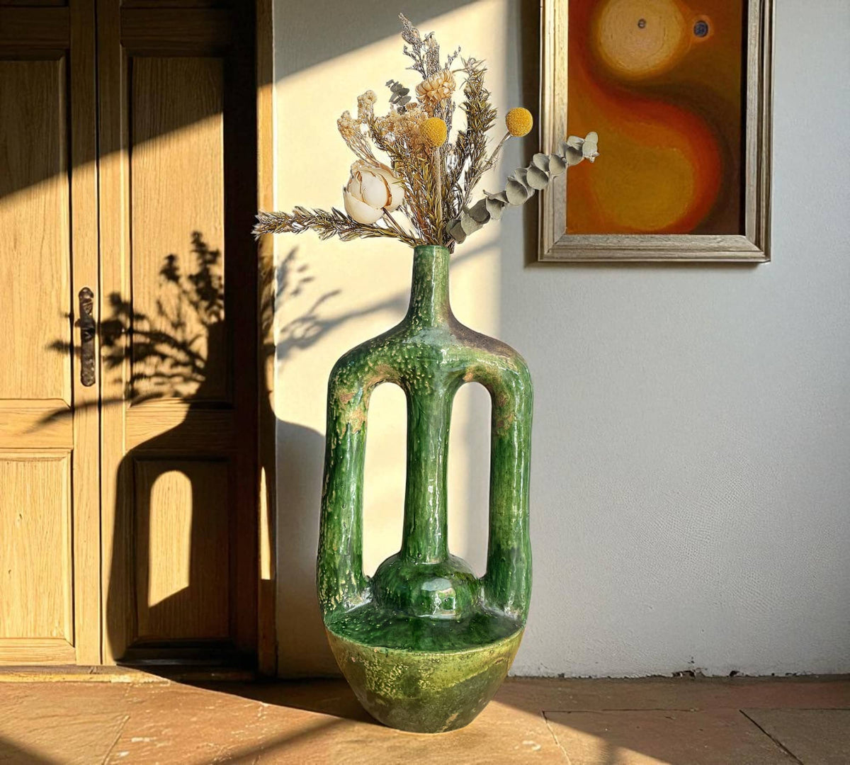 Green ceramic vase with dried flowers against a wooden door and wall art.
