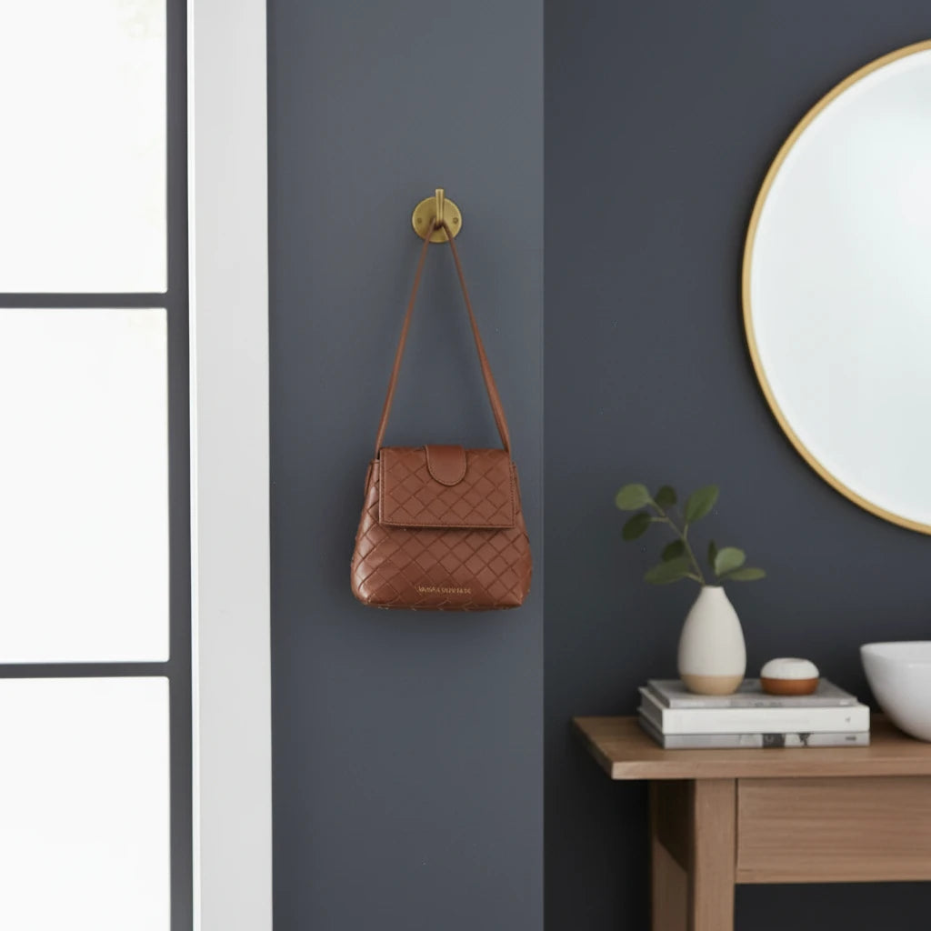 Brown quilted handbag hanging on a wall hook next to a round mirror and wooden side table with decorative items.