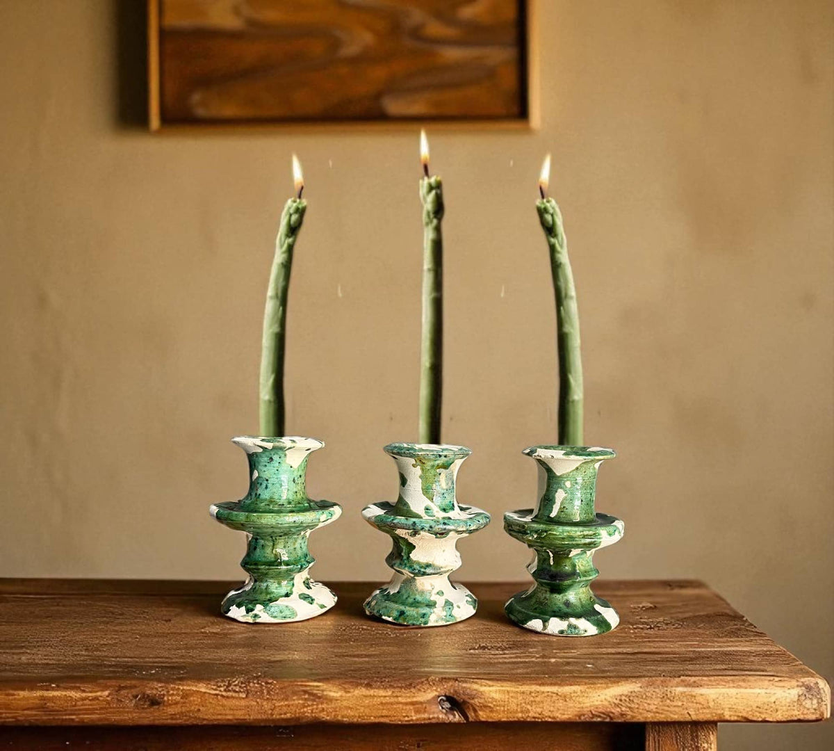 Three green ceramic candle holders with lit candles on a wooden surface against a beige wall.