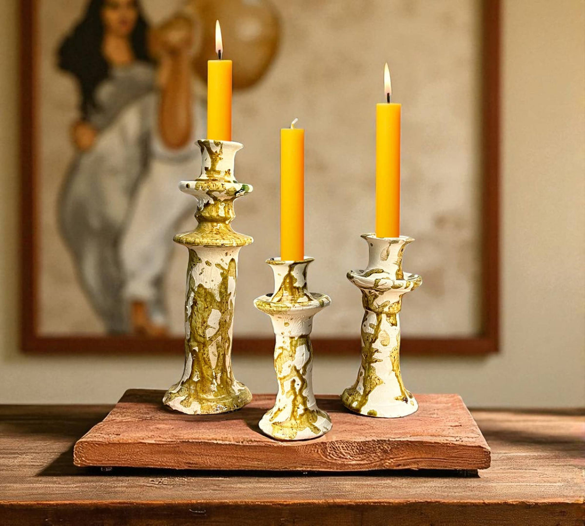 Three candle holders with candles on a wooden stand in front of a painting.