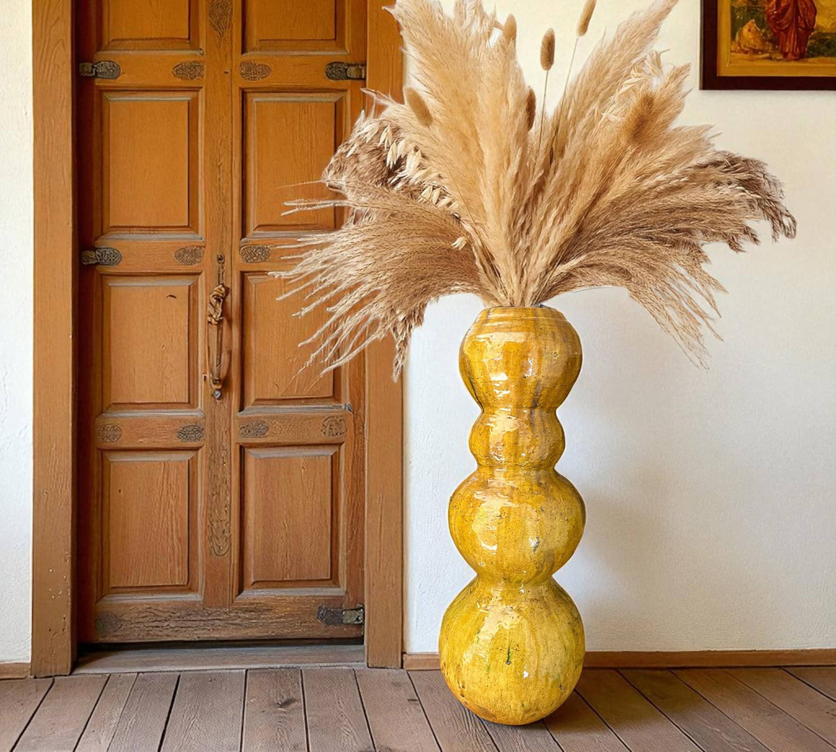 Yellow vase with dried pampas grass against a wooden door and white wall.