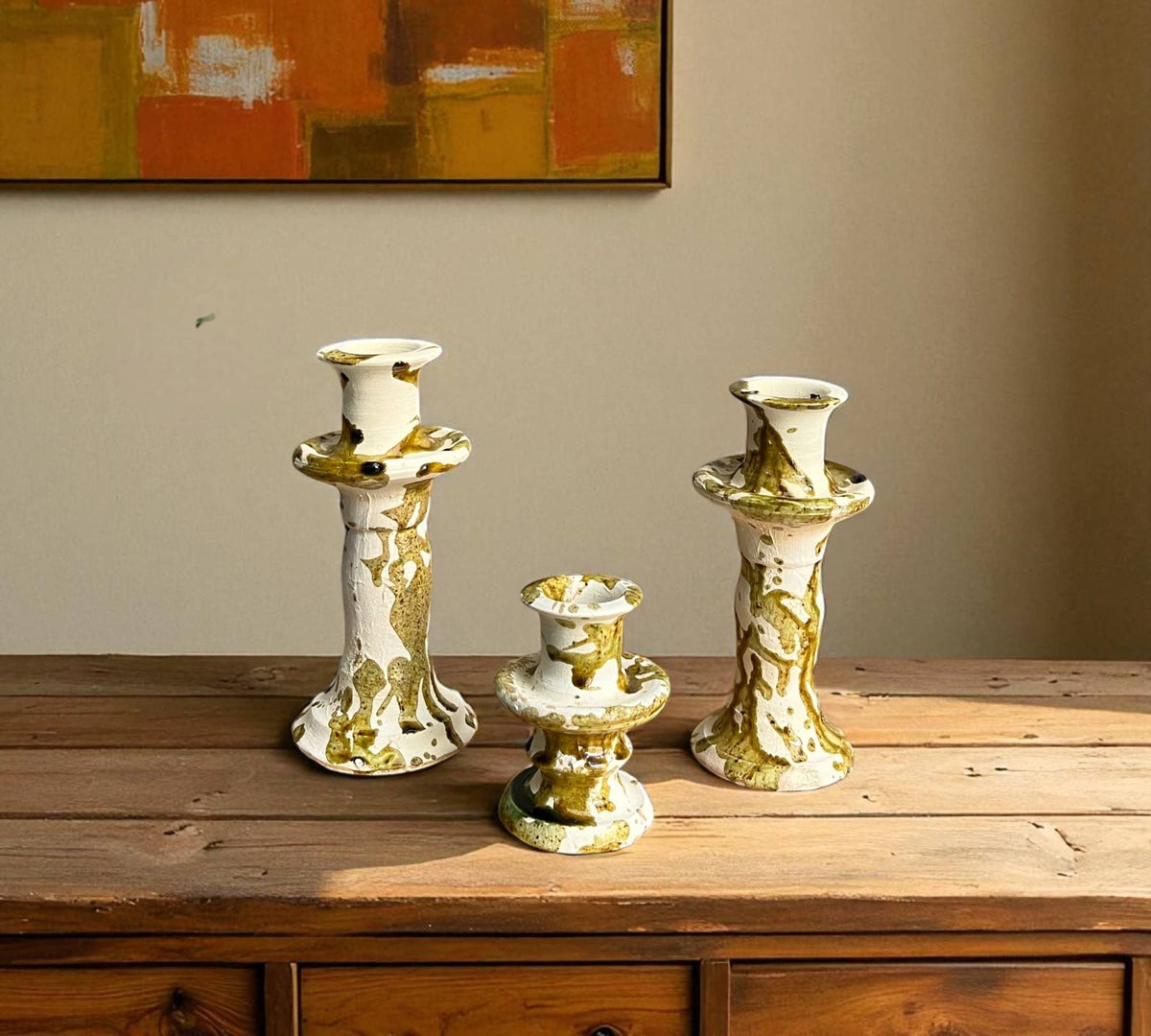 Three decorative candle holders on a wooden surface with a painting in the background.