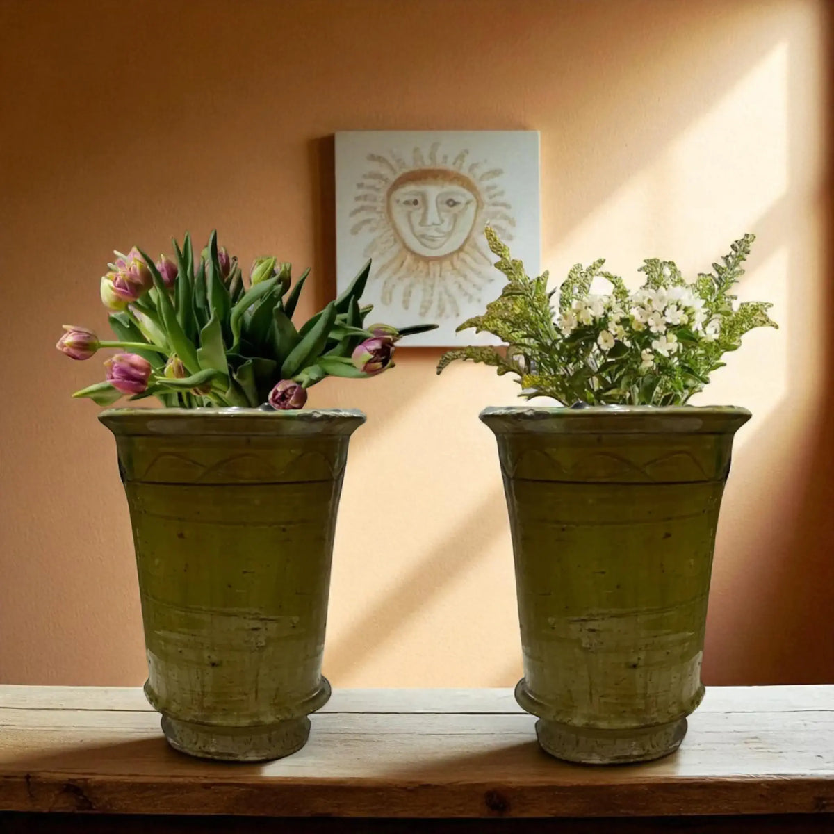 Two rustic yellow pots with plants on a wooden surface against an orange wall with a sun-themed painting.