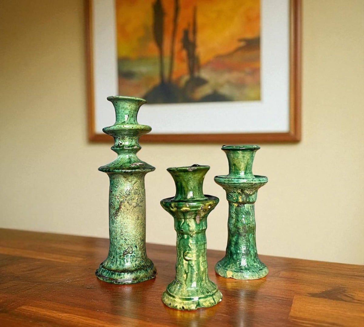 Three green ceramic candle holders on a wooden surface with a painting in the background.