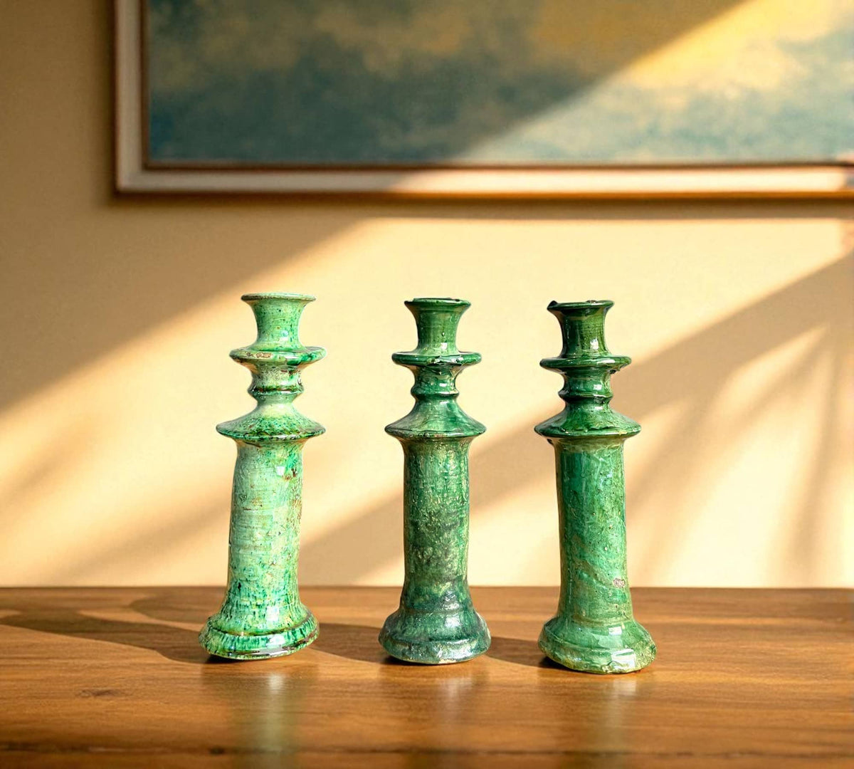 Three green ceramic candle holders on a wooden surface with a painting in the background.