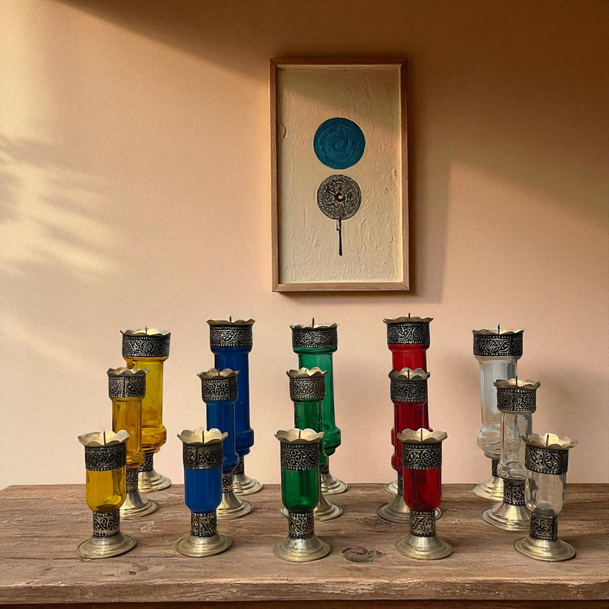 Vintage hand‑engraved silver‑plated brass and recycled glass candlesticks in assorted colours on a wooden surface with a framed artwork in the background.