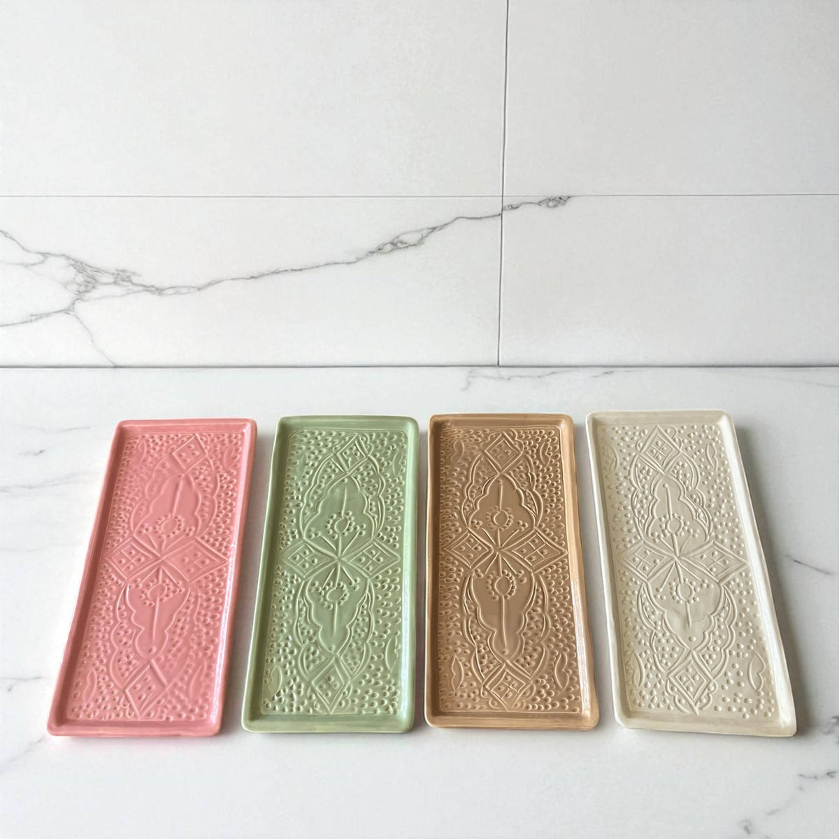 Four rectangular ceramic trays with embossed designs in pink, green, beige, and cream on a marble surface.