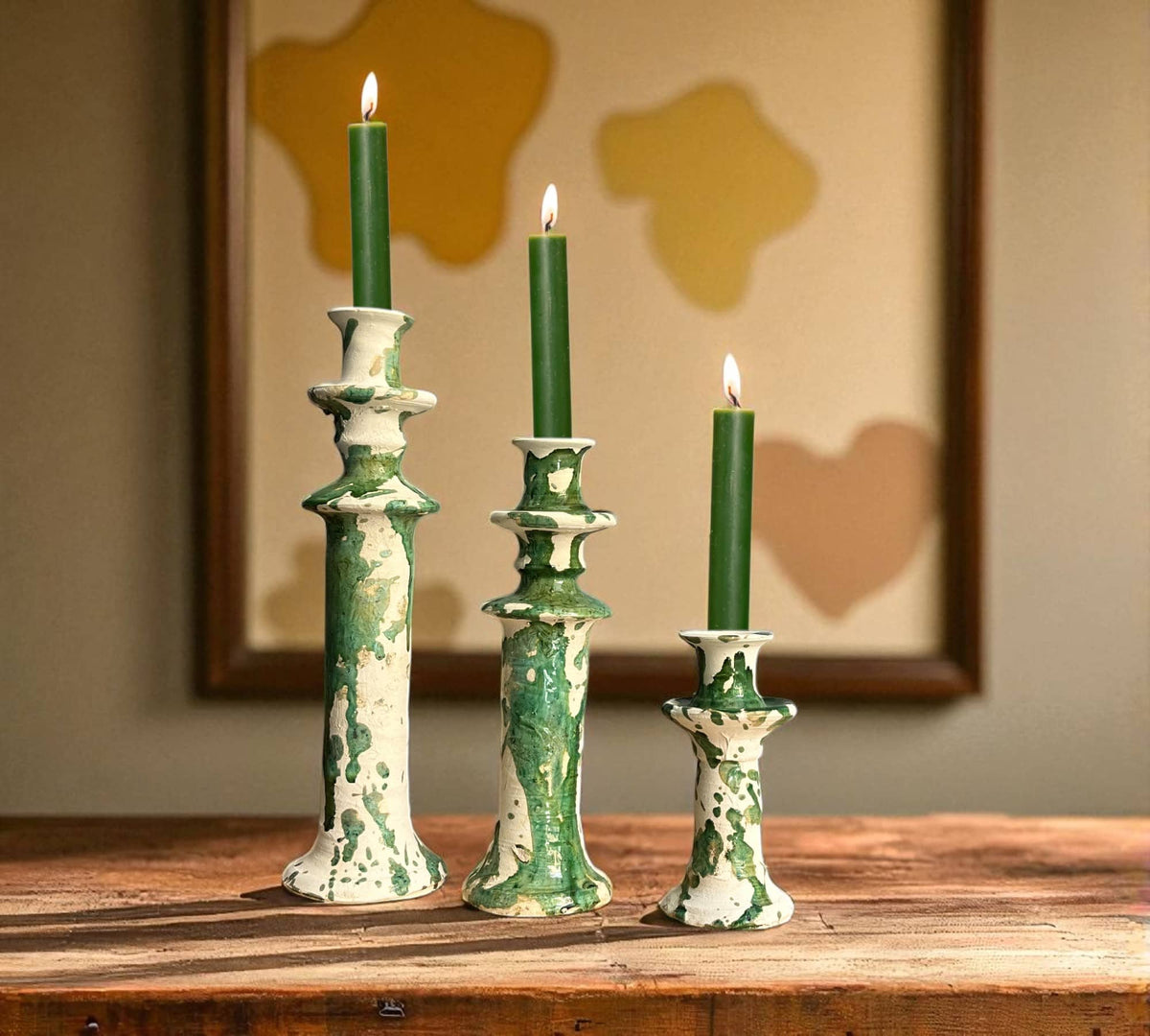Three green candles in decorative candlesticks on a wooden surface with a blurred abstract painting in the background.