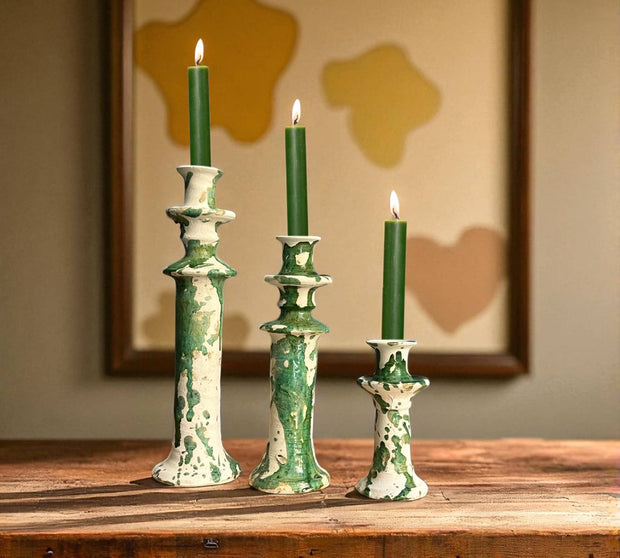 Three green candles in decorative candlesticks on a wooden surface with a blurred abstract painting in the background.