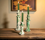 Decorative candlesticks with green candles on a wooden surface, with a blurred painting in the background.