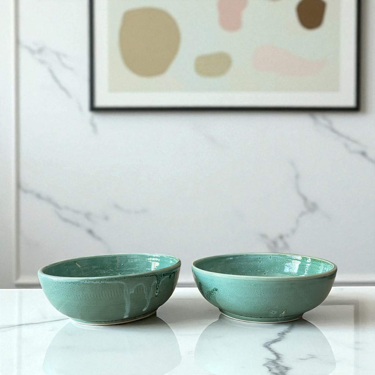 Two green ceramic bowls on a marble surface with abstract art in the background.