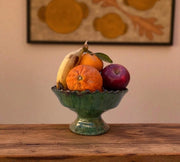 Fruit bowl with oranges, bananas, and a plum on a wooden surface with a decorative wall hanging in the background.