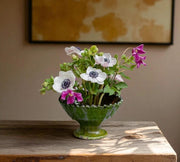 Green vase with flowers on a wooden surface against a beige wall.