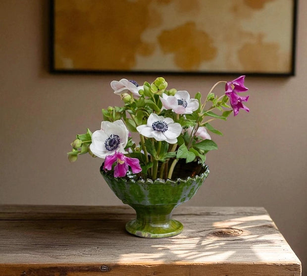 Green vase with flowers on a wooden surface against a beige wall.