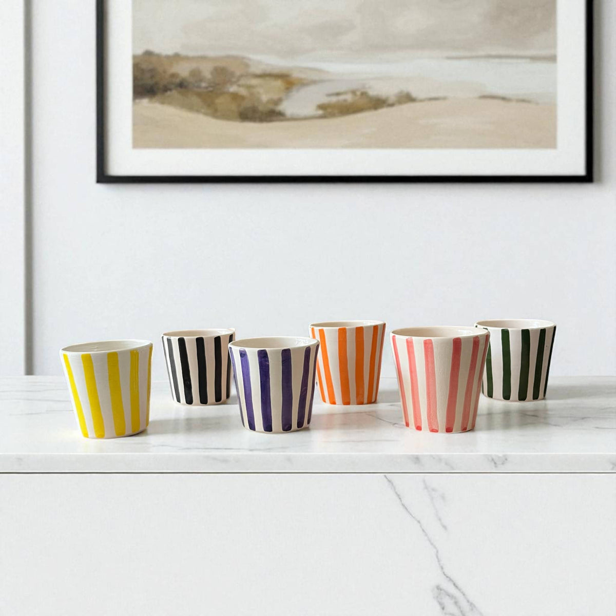 Set of colorful striped cups on a white surface with a framed landscape painting in the background.