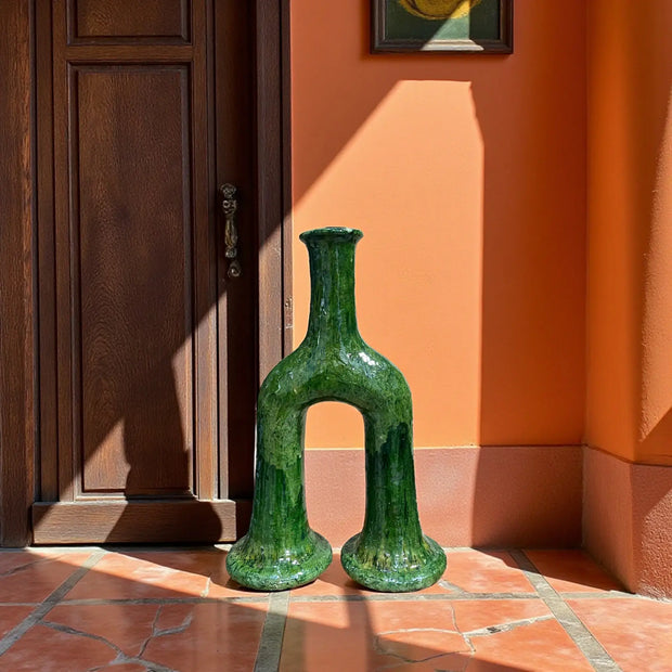 Green ceramic vase on a tiled floor with an orange wall and wooden door in the background