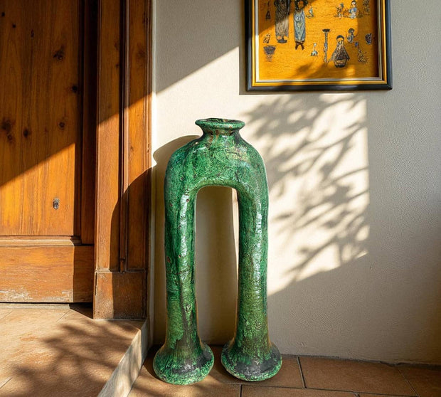 Green ceramic vase on a tiled floor with wooden paneling and framed artwork in the background.