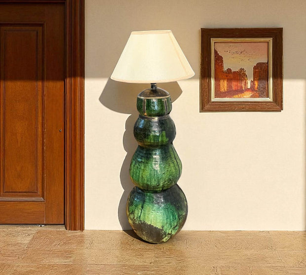 Green ceramic floor lamp with a white shade in a room with a wooden door and framed picture on the wall.