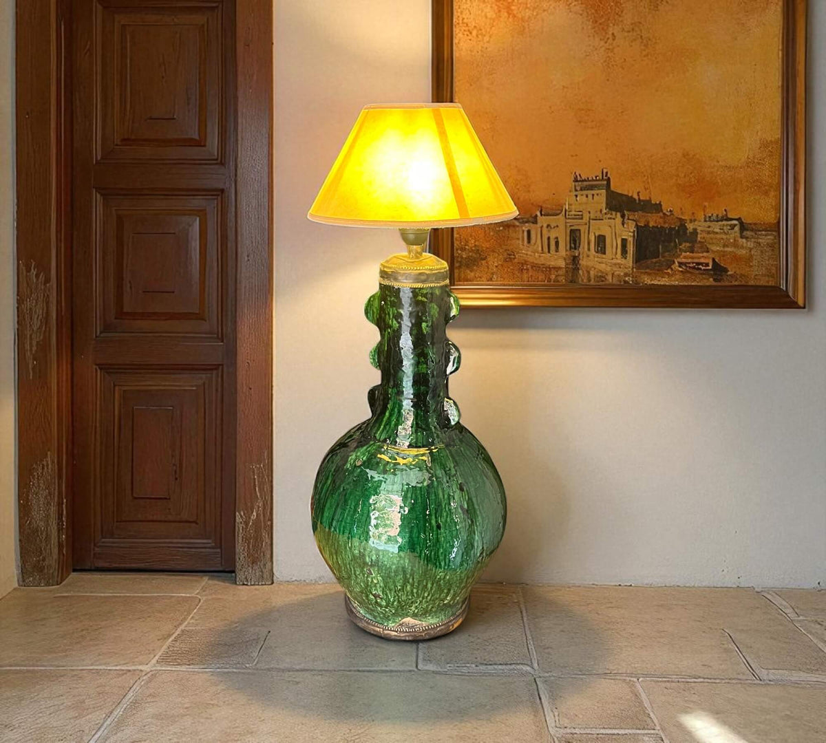 Green ceramic floor lamp with yellow lampshade in a room with a wooden door and framed artwork.