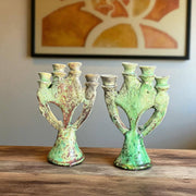 Two green ceramic candle holders on a wooden surface with a framed abstract painting in the background.