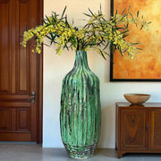 Green textured vase with yellow flowers in a room with wooden furniture and a painting.