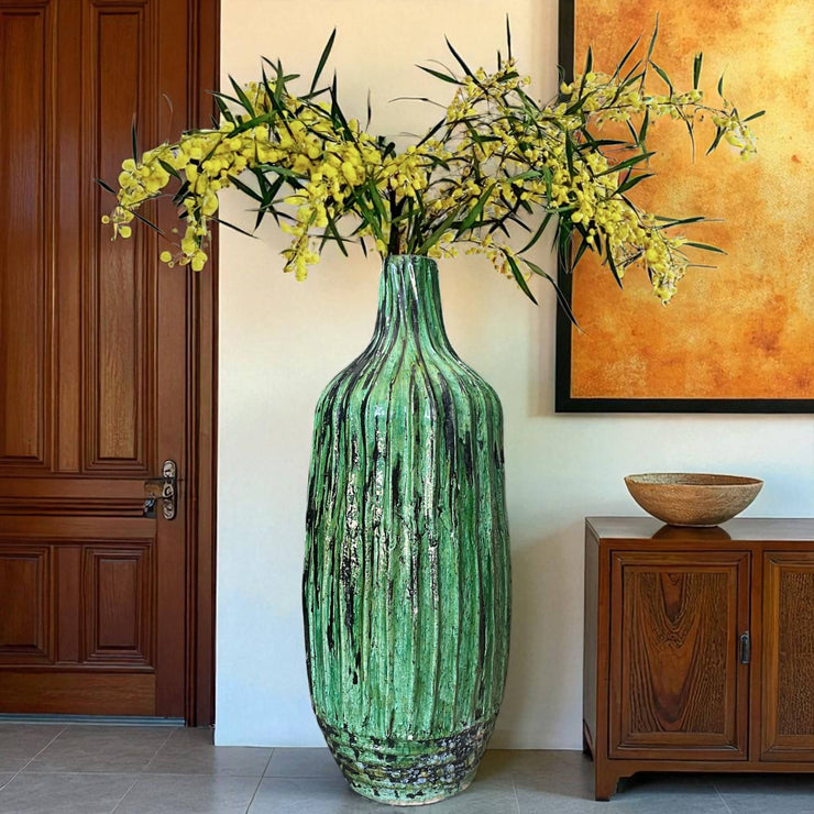 Green textured vase with yellow flowers in a room with wooden furniture and a painting.