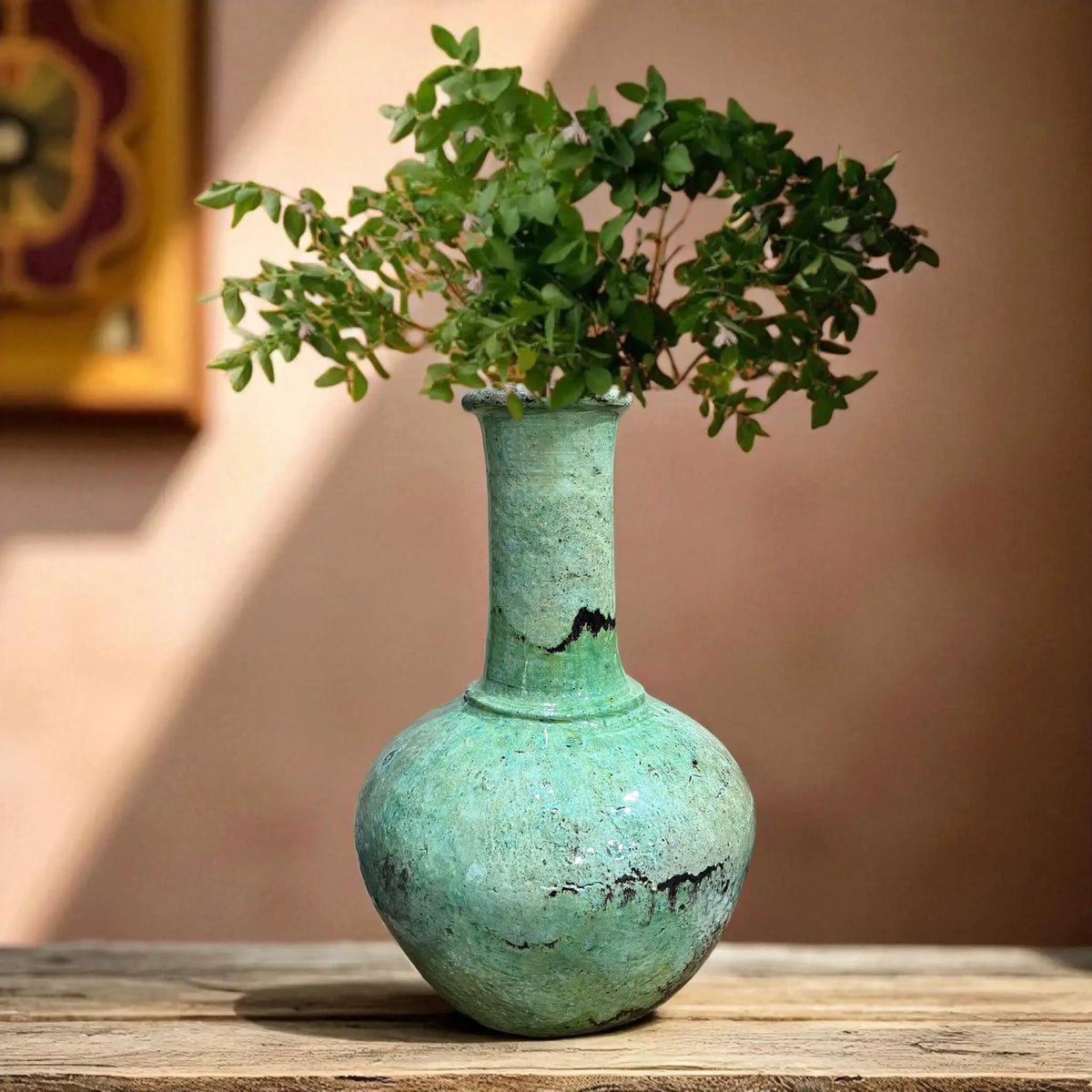 Vintage green vase with a plant on a wooden surface against a beige wall.