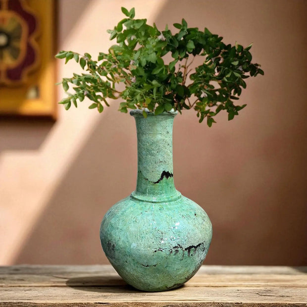 Vintage green vase with a plant on a wooden surface against a beige wall.