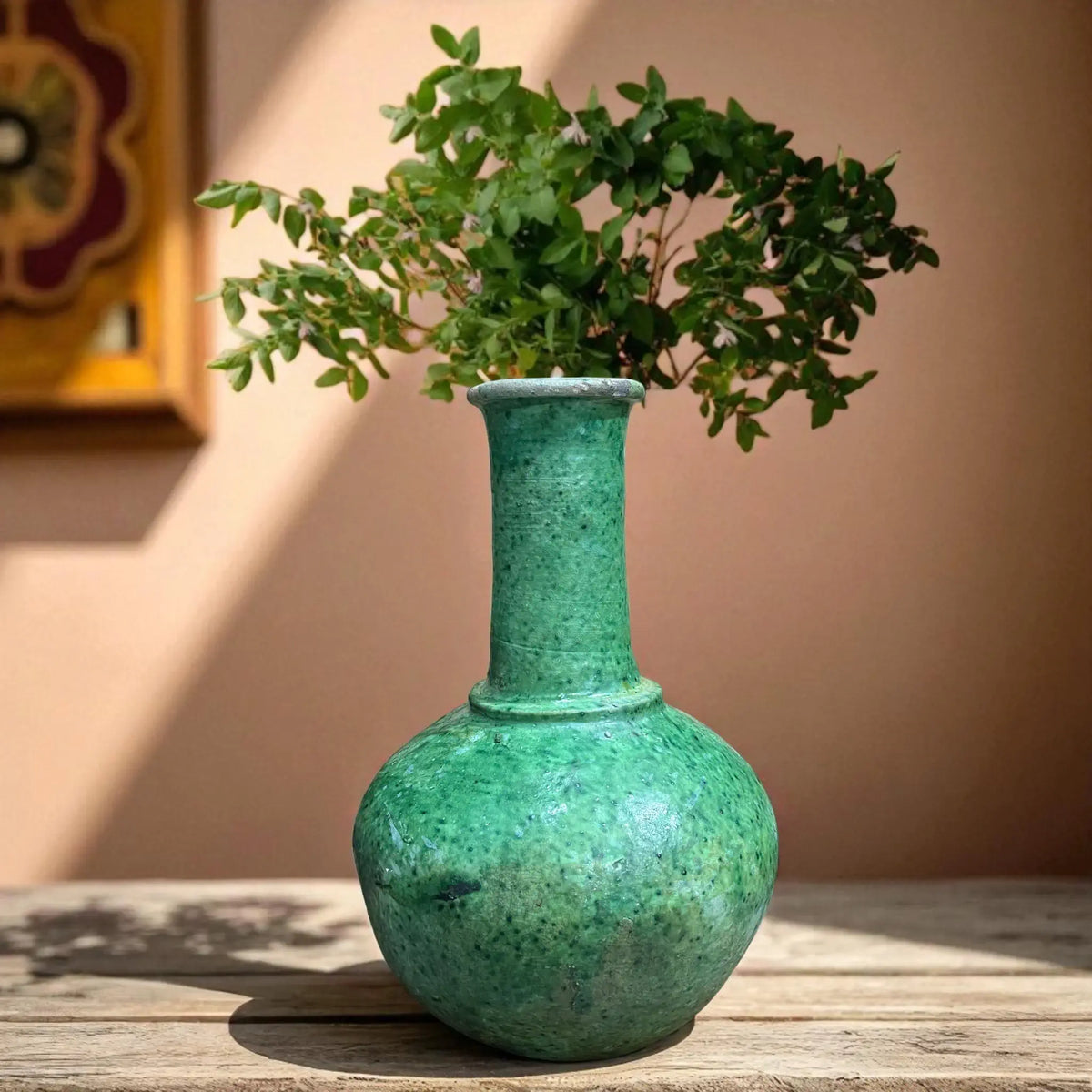 Green textured vase with a plant on a wooden surface against a beige wall.