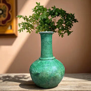 Green textured vase with a plant on a wooden surface against a beige wall.