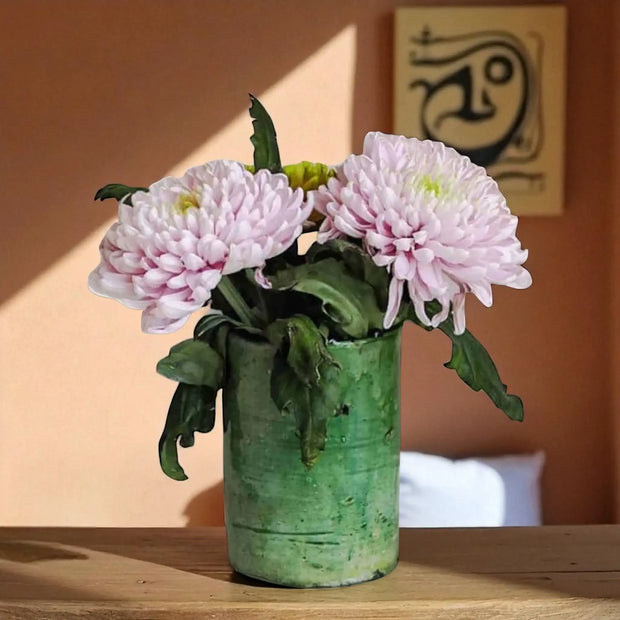 Pink flowers in a green vase on a wooden surface with a warm-toned wall in the background.