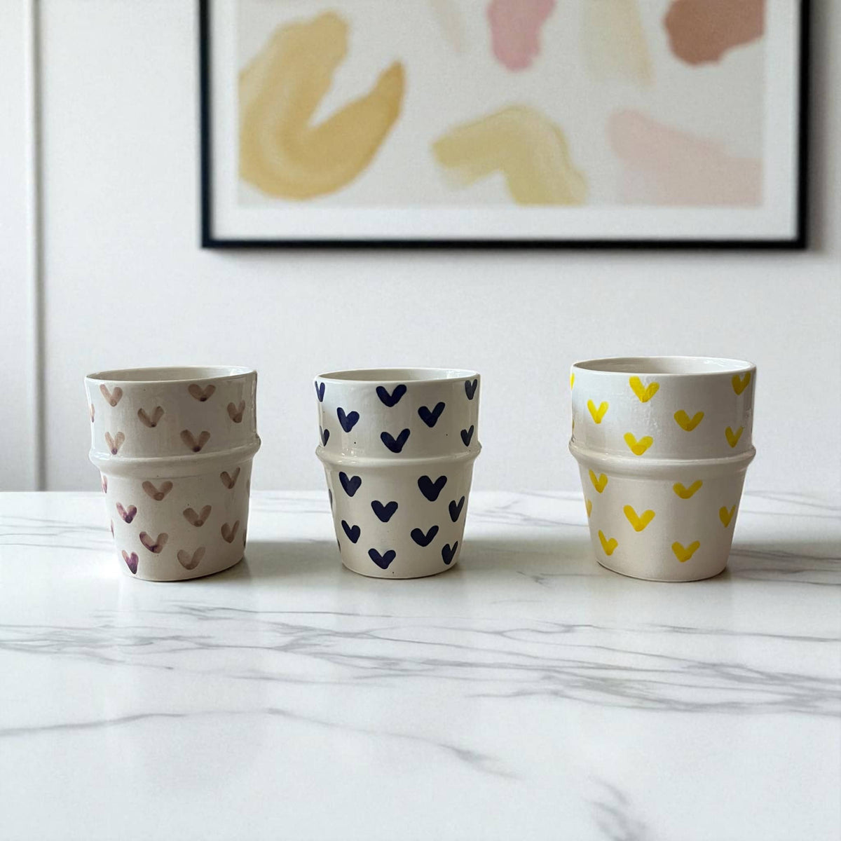 Three stacked ceramic pots with heart patterns on a marble surface, framed abstract art in the background.