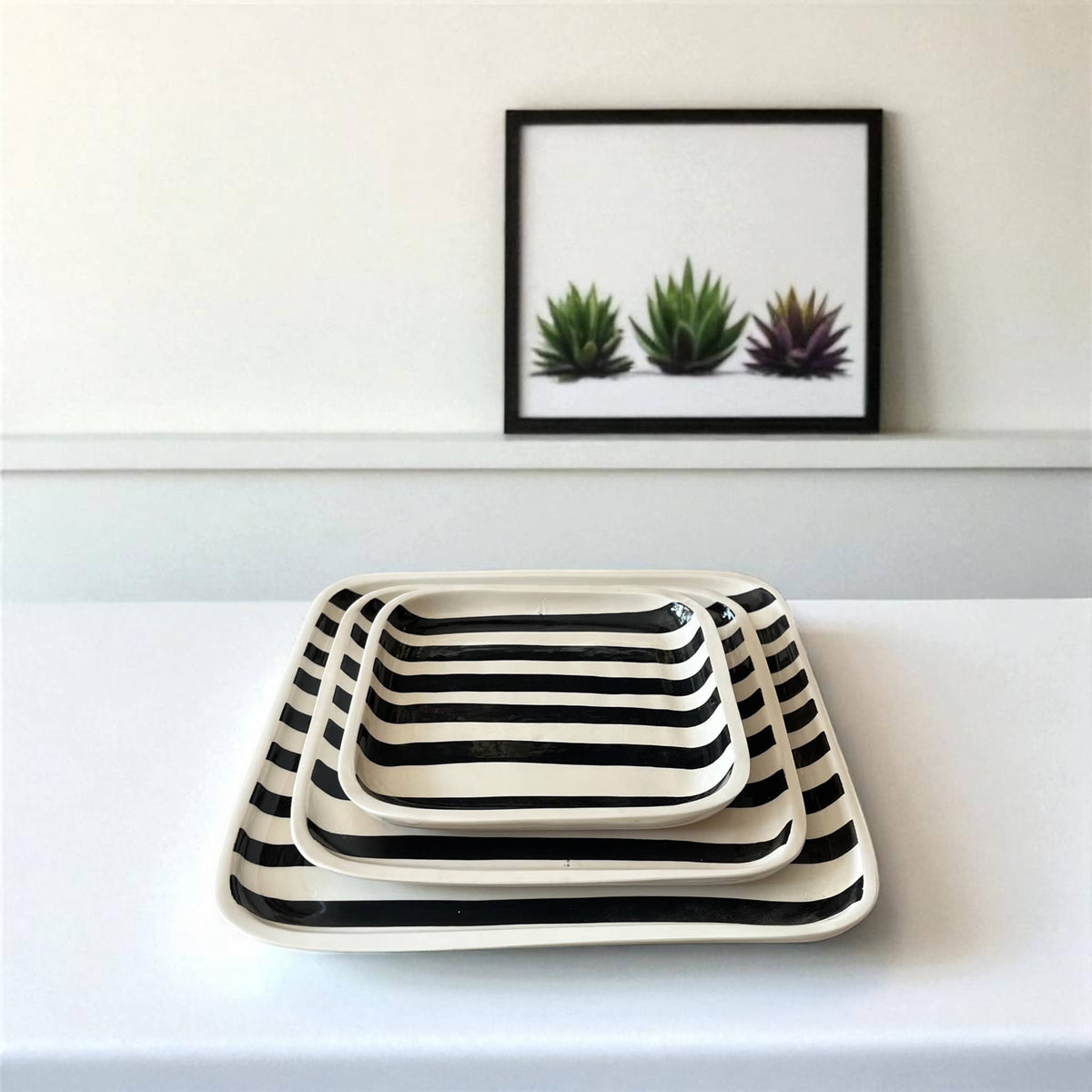 Set of three black and white striped trays on a white surface with a framed picture of plants in the background.