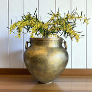 Decorative brass vase with yellow flowers on a wooden surface against a white paneled wall.