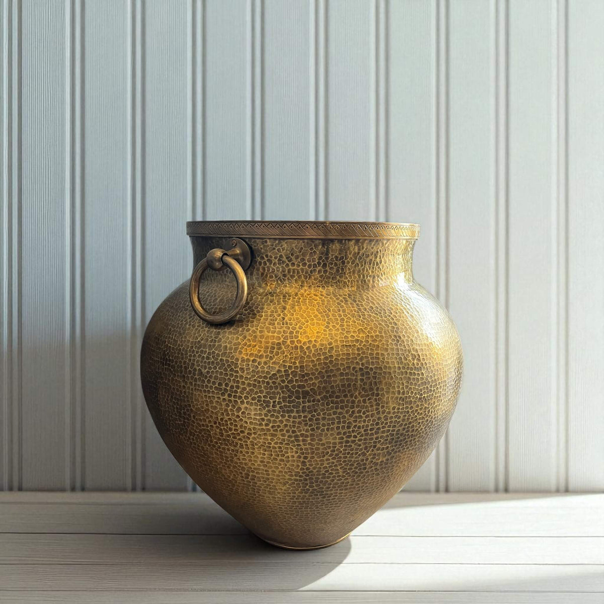 Bronze vase with textured surface and circular handle on a light wooden surface with a white paneled wall background.