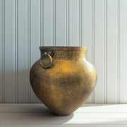 Bronze vase with textured surface and circular handle on a light wooden surface with a white paneled wall background.