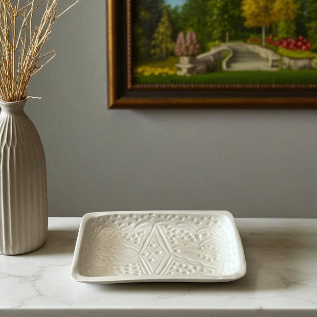 White ceramic tray with geometric pattern on a marble surface, next to a vase with dried plants and a framed landscape painting.